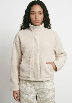 Une femme porte une veste sherpa couleur crème à col montant et poches, assortie à un pantalon camouflage clair, sur un fond uni.