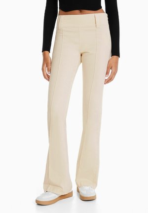 Vrouw die beige hoge taille uitlopende broek en witte sneakers draagt, getoond van taille tot voeten tegen een effen achtergrond.