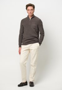 Brauner, gerippter Strickpullover mit einem halben Reißverschlusskragen und einer Fronttasche, kombiniert mit hellcremigen Hosen und schwarzen Schuhen.