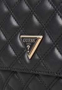 Schwarze, gesteppte Tasche aus Kunstleder mit einem goldenen, dreieckigen Logo-Plakette und schwarzen Akzenten, genähten Details und texturierten Oberflächen.