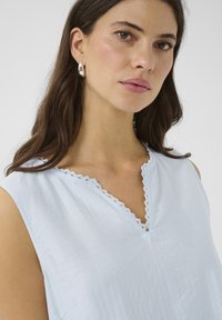 Blouse bleu clair en tissu texturé, avec un décolleté en V bordé de dentelle, sans manches, affichant une coupe décontractée.