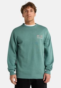 Sweatshirt vert avec col rond, présentant des poignets et un ourlet côtelés, orné d'un petit logo multicolore Billabong sur le côté gauche de la poitrine. Tissu doux et texturé.