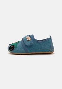 Modrá textilná papuča so zeleným dizajnom traktora na špičke, s lepiacim pásikom a gumovou podrážkou. Textúrovaný materiál pripomínajúci denim.