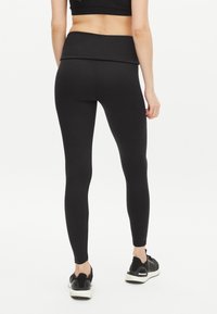 Kvinna iklädd svarta högmidjade leggings och svarta sneakers, stående med ryggen mot kameran mot en vit bakgrund.