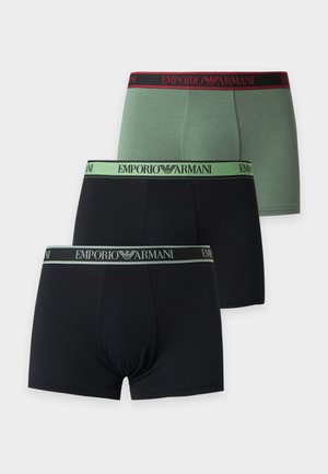 Mladý muž stojaci naboso, nosiaci čierne boxerky Emporio Armani so svetlozeleným pásom, na bielom jednoduchom pozadí.