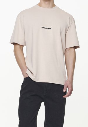 Beżowy, krótkim rękawem t-shirt wykonany z miękkiej tkaniny, z subtelnym czarnym logo "PEGADOR" na piersi i luźnym krojem.