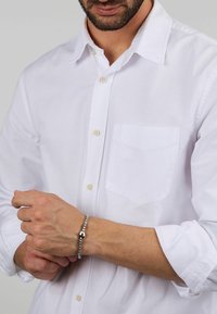Camicia bianca a maniche lunghe con un'unica tasca sul petto, maniche arrotolate fino al gomito, indossata con un braccialetto d'argento al polso.