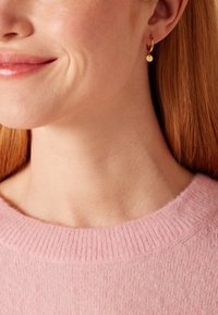 Des boucles d'oreilles en or en forme de créneau présentent un petit charme rond, porté sur un cou avec un pull texturé rose clair. L'ensemble du look est minimaliste.