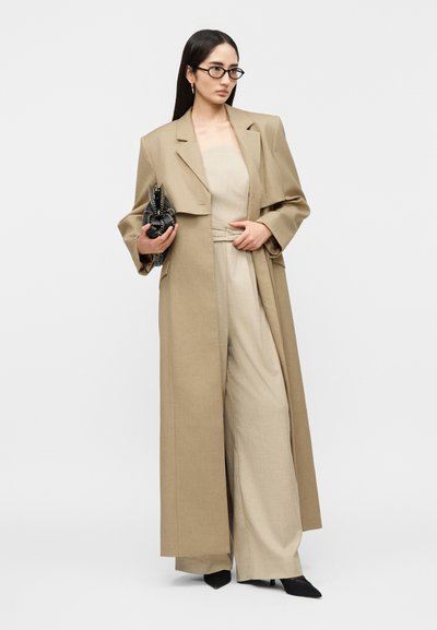 Γυναίκα με μακριά σκούρα μαλλιά που φοράει μπεζ καμπαρντίνα και φαρδιά παντελόνια, κρατώντας μαύρο πλεκτό τσαντάκι, φοράει μαύρα τακούνια και γυαλιά.