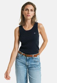 Navyblauw mouwloos gebreid tanktop met kabelpatroon en ronde hals; gedragen met hoog-waisted lichtblauwe jeans en een bruine riem.