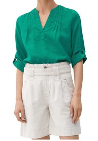 Grüne Leinenbluse mit V-Ausschnitt und Falten-Detail, kombiniert mit cremefarbenen hochtaillierten Shorts mit einem breiten Gürtel und seitlichen Taschen.