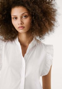 White shirt with ruffled short sleeves, featuring a button-down front and a textured finish. Smooth fabric and modern design.