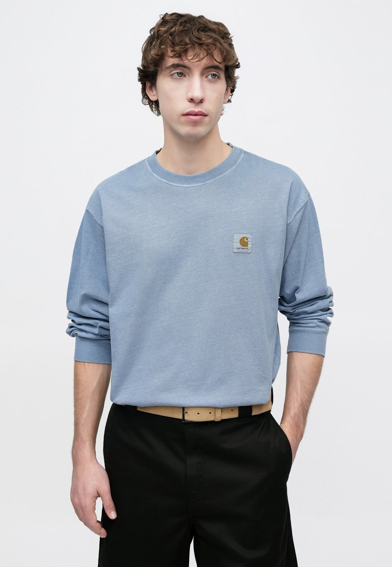 Felpa azzurro chiaro a maniche lunghe con collo rotondo e polsini arrotolati, con una toppa con logo quadrata sul petto, abbinata a pantaloni neri.