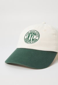 Cremefarbener Cap mit grünem Schild und gesticktem "NYC Athletic Club"-Logo in einem kreisförmigen Design. Strukturierter Stoff, verstellbarer Hinterriemen.
