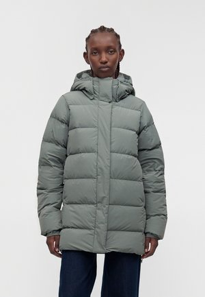 Personne portant une doudoune d’hiver à capuche vert clair, les mains détendues le long du corps, sur un fond gris uni.