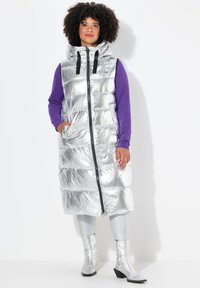 Langer silberner Daunenweste mit Kapuze, mit horizontalen Nähten, vorderem Reißverschluss und schwarzen Akzenten. Kombiniert mit metallischen Leggings und Stiefeln.