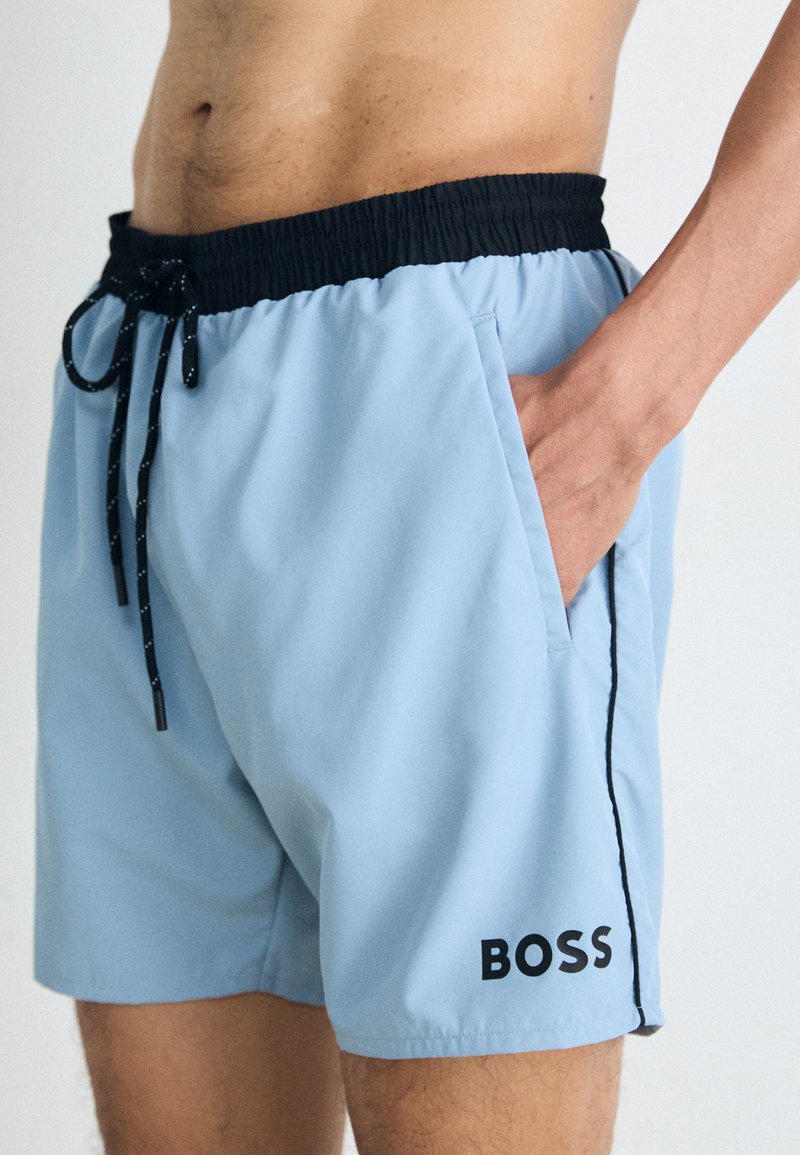 Shorts de bain bleu clair avec une taille élastique noire, un cordon de serrage, des poches latérales et un logo "BOSS" noir en bas à gauche.