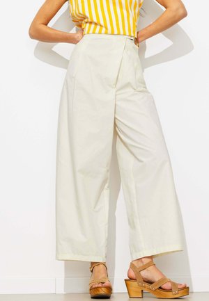 Femme portant un pantalon crème taille haute jambes larges, un top sans manches rayé jaune et blanc, et des sandales compensées marron avec des brides à la cheville.
