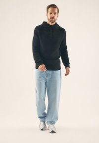 Pull à capuche noir en maille avec col châle, associé à un jean ample bleu clair et des chaussures de sport argentées. Le mannequin pose devant un fond neutre.