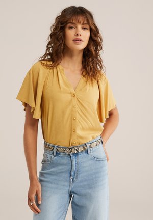 Vrouw met golvend bruin haar die een gele blouse met knopen en vliegermouwen draagt, lichtblauwe spijkerbroek en een riem met slangenprint.