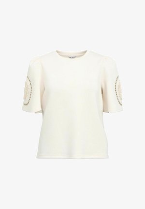 Crème-kleurige top met korte mouwen en pofmouwen, met een cirkelvormig structuurpatroon op de linker mouw en een ronde halslijn.