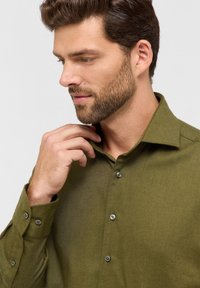 Camicia verde con bottoni e tessuto texture. Presenta un colletto a punta e bottoni, mostrata mentre viene aggiustata al collo.