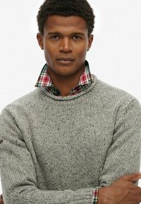 Suéter gris de punto con cuello redondo, que presenta un cuello de cuadros en rojo, verde y blanco. La textura parece áspera con motas.