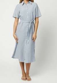 Donna che indossa un abito midi a righe blu chiaro e bianche con bottoni, maniche corte, cintura in vita annodata e ciabatte bianche.