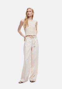 Top corto de color beige claro con bordes texturizados y con volantes, combinado con pantalones amplios de tiro alto en color crema. Chanclas negras completan el look.
