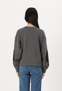Felpa grigia dal taglio rilassato, con scollo rotondo e ricamo decorativo nero sulla manica sinistra. Indossata con jeans blu.