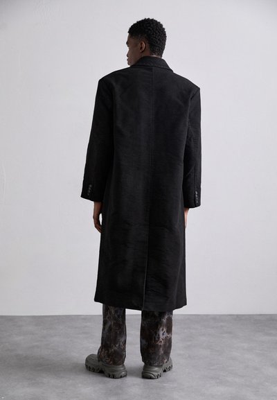Long, black coat made of textured fabric, featuring a classic collar and a straight cut. Worn with patterned trousers and bulky shoes.