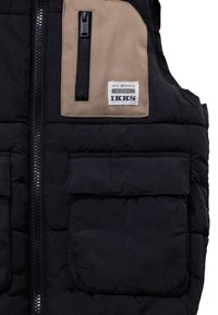 Chaleco acolchado negro con bolsillo en el pecho beige y cremallera negra, que presenta una etiqueta blanca de IKKS Heritage y un bolsillo adicional con tapa en la parte frontal.