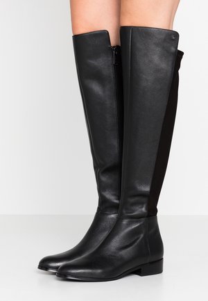 Sorte læderstøvler til knæene med lave hæle og en lynlås på siden, båret på bare ben foran en neutral hvid baggrund.