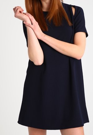 Femme portant une robe noire à manches courtes avec des découpes aux épaules, tenant son poignet gauche avec sa main droite.