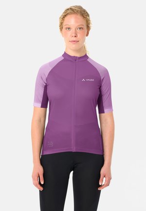 Donna con capelli biondi ricci, indossa una maglia da ciclismo viola a maniche corte e pantaloncini da ciclismo neri, in piedi rivolta verso di fronte.