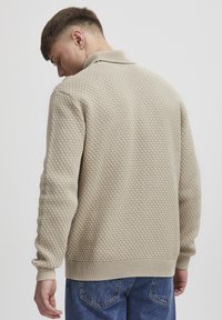 Beige textured sweater with a honeycomb pattern, ribbed collar and cuffs, and a relaxed fit. Worn with blue jeans, showing the back view.