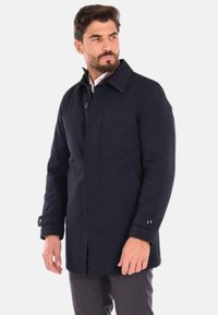 Cappotto blu navy con colletto abbottonato, tasche frontali a fessura e polsini con bottoni, realizzato in un tessuto liscio e testurizzato.