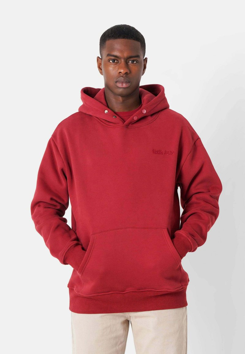 Sudadera de algodón roja con un bolsillo frontal, capucha ajustable con cordón y puños de canalé. Presenta un texto sutil en el pecho en un color a juego.