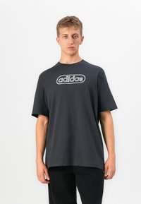 Νέος άνδρας με κοντά μαλλιά, φορώντας ένα χαλαρό μαύρο μπλουζάκι Adidas και μαύρο παντελόνι, στέκεται μπροστά σε μια λευκή επίπεδη επιφάνεια.