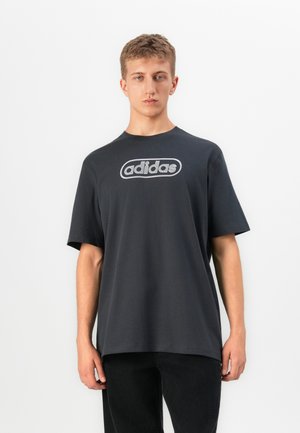 Young man with short hair wearing a loose black Adidas t-shirt and black pants standing against a plain white background.