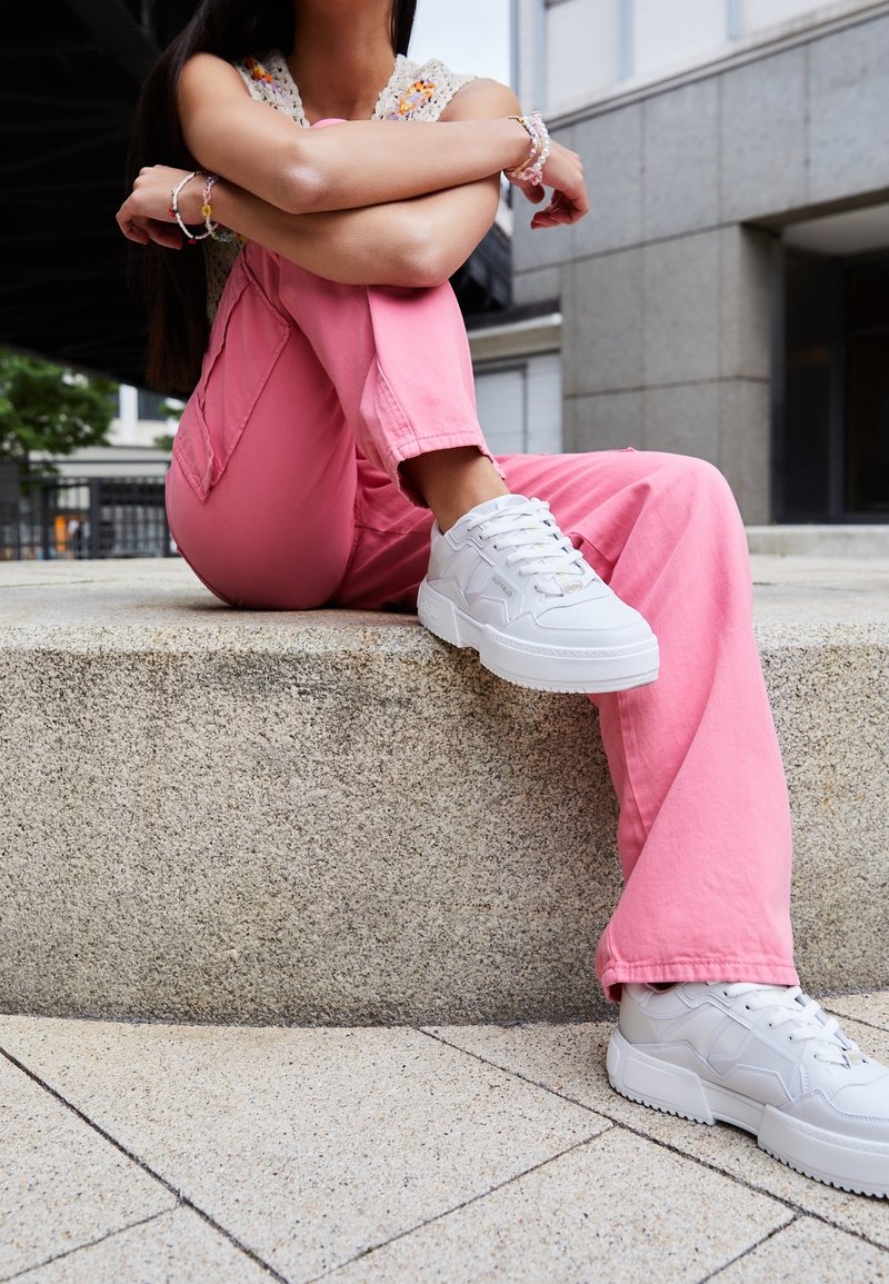 Vita sneakers med texturerad detaljering, kombinerade med rosa tygbyxor. Szenen har en stenplattform och en urban miljö.