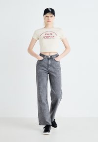 Camiseta recortada crema con texto gráfico rojo, combinada con jeans de mezclilla gris de cintura alta con acentos de tachuelas plateadas. Zapatillas negras y gorra.