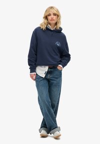 Donna bionda che indossa un felpa blu scuro, una maglietta bianca sotto, jeans a gamba larga con risvolto e sneakers bianche, in piedi con una mano in tasca.