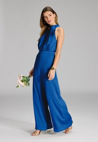 Tuta blu con scollatura a halter e dettaglio a fiocco, gambe larghe, texture a costine, abbinato a tacchi dorati e mentre tiene un bouquet di fiori.