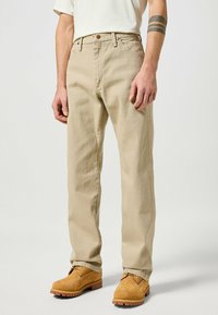 Beige cotton trousers with a straight-leg design, featuring front pockets and a brown button. Worn with light brown lace-up boots.
