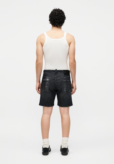 Άνδρας με σγουρά μαλλιά που φοράει λευκό αμάνικο μπλουζάκι, μαύρα σκισμένα τζιν σορτς, λευκές κάλτσες και μαύρα παπούτσια, στέκεται γυρισμένος με την πλάτη.