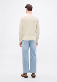 Béžový pletený sveter s dlhými rukávmi, skombinovaný so svetlomodrými džínsami s širokými nohavicami. Semišové hnedé topánky. Jednoduché pozadie.