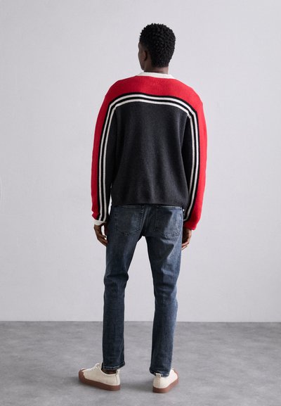 Maglione a strisce nero e rosse con corpo grigio, dotato di collo rotondo e polsini a costine, abbinato a jeans blu e sneakers beige.