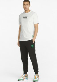 Tricou alb din bumbac cu logo-ul negru "PUMA", asortat cu pantaloni de trening negri cu accente verzi și logo. Încălțări colorate completează ținuta.