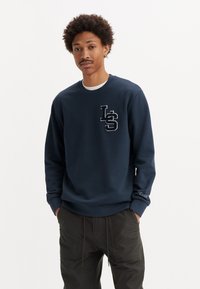 Jonge man met een afro kapsel, die een marineblauwe sweatshirt draagt met een "LS" logo op de borst en donkere broek, met handen in de zakken, op een neutrale achtergrond.